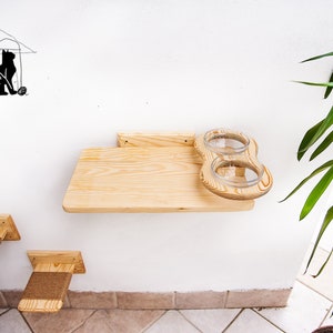 Op de afbeelding: Een houten kattenvoederstation met twee glazen kommen gemonteerd op een plank. De plank is bevestigd aan een witte muur. Er zijn twee kleinere houten platforms met touw bedekte oppervlakken onder de plank.