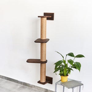May include: A brown wooden cat scratching post with three platforms and sisal rope covered posts. The scratching post is mounted to a white wall. A green plant in a yellow pot is in the foreground.