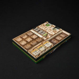 May include: A wooden game organizer with multiple compartments, designed to hold game cards and components. The organizer has a green and gold trim and is filled with various game pieces. The text "Caverna Laserox" is visible on the top.