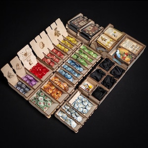May include: Wooden game organizer with multiple compartments filled with colorful game pieces, cards, and small black pouches. The organizer is made of light-colored wood and features laser-cut designs and text.
