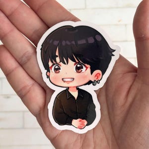 May include: A sticker featuring a cartoon illustration of a person with black hair, brown eyes, and a black shirt. The character has a small smile and is wearing earrings. The sticker is held in a hand.