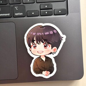 May include: A sticker featuring a cartoon illustration of a person with dark hair and a black shirt. The character has red eyes and is smiling. The sticker is placed on a laptop keyboard.