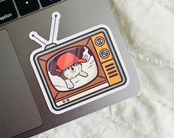 Pode incluir: Um adesivo de um cão de desenho animado usando um boné de beisebol vermelho e deitado em um travesseiro dentro de uma televisão vintage.