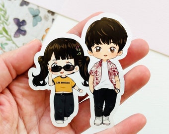 May include: Two cartoon stickers of a girl and a boy. The girl is wearing a yellow shirt with the words "Los Angeles" on it, black pants, and sunglasses. The boy is wearing a red and white patterned shirt, a white undershirt, black pants, and white shoes.