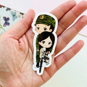 May include: A sticker featuring a cartoon couple riding a bicycle. The man is wearing a camouflage uniform and a hat with a star. The woman is wearing a pink dress and has long black hair. The sticker is on a white background.