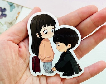 May include: A sticker featuring a cartoon couple. The woman is wearing a pink sweater and has long black hair. The man is wearing a black jacket and has short black hair. They are both looking down with sad expressions. The woman is holding a red suitcase.