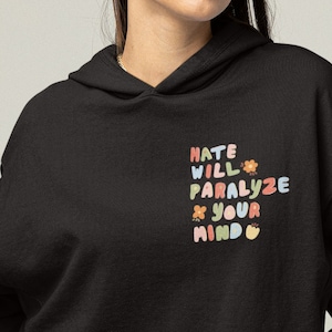 Puede incluir: Sudadera con capucha negra con un estampado gráfico que dice "Hate Will Paralyze Your Mind" en letras de colores con flores.