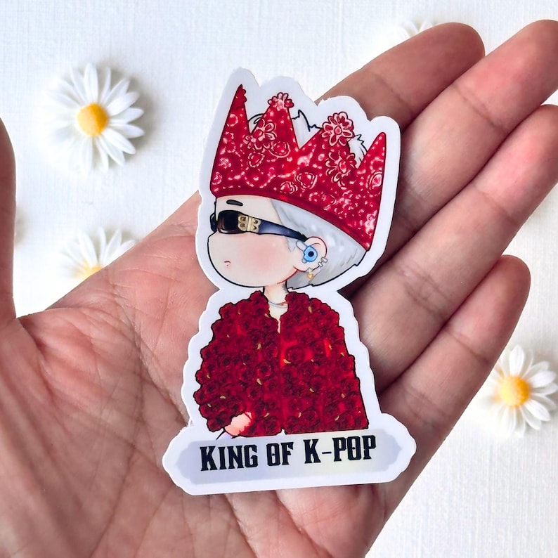 Puede incluir: Una pegatina con un personaje de dibujos animados con una corona floral roja, gafas de sol y una chaqueta floral roja. El texto "KING OF K-POP" se muestra en una pancarta blanca. Las margaritas blancas est&aacute;n en el fondo.
