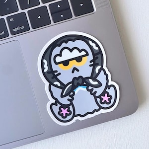 May include: A die-cut sticker featuring a cartoon character with a grumpy expression. The character is primarily gray with black and white accents, yellow eyes, and pink star-shaped feet. The sticker is placed on a laptop.