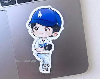 May include: A sticker featuring a cartoon illustration of a person wearing a blue baseball cap with the Los Angeles Dodgers logo. The person is wearing a white jersey with "Dodgers" printed on it, light blue pants, and a black glove. The sticker is on a laptop.