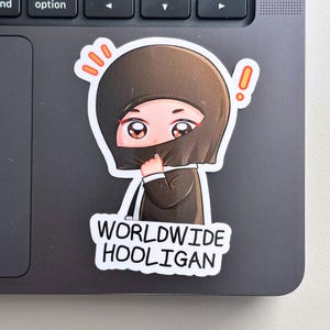 Peut inclure: Un autocollant représentant un personnage de dessin animé avec une capuche marron, cachant partiellement son visage. Le texte "WORLDWIDE HOOLIGAN" est en blanc. Le personnage a de grands yeux et est sur un ordinateur portable.