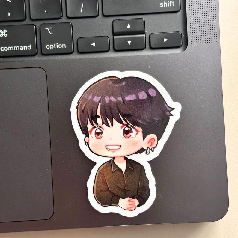 May include: A sticker featuring a cartoon illustration of a person with dark hair and a black shirt. The character has red eyes and is smiling. The sticker is placed on a laptop keyboard.