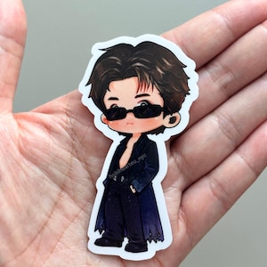 May include: A die-cut vinyl sticker featuring a stylized cartoon illustration of a person with dark brown hair wearing black sunglasses, a dark open blazer, and dark trousers. The text hobihearteu_nyc is printed in small white letters on the blazer.