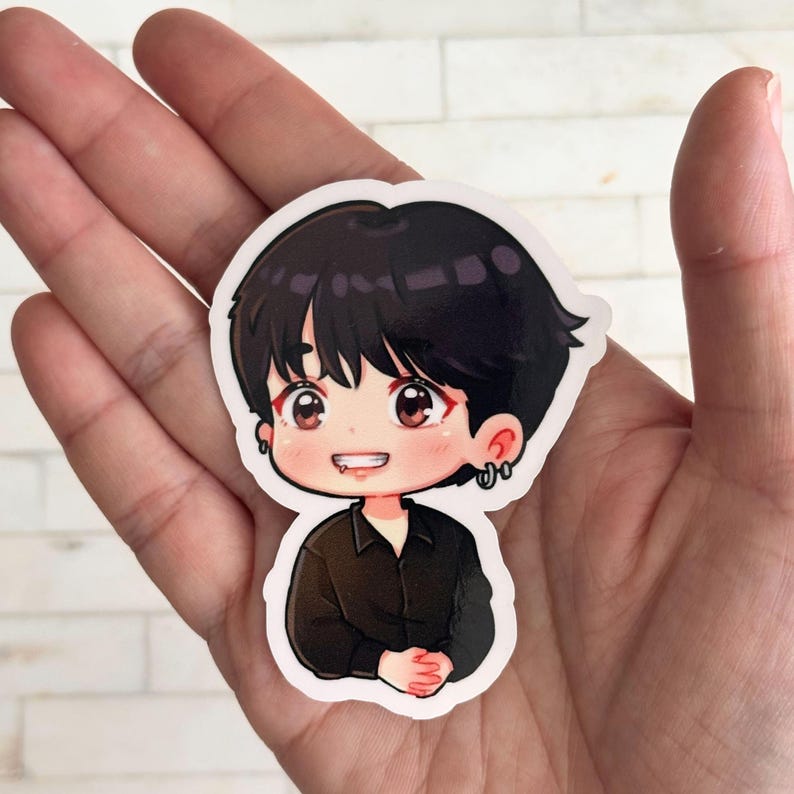 May include: A sticker featuring a cartoon illustration of a person with black hair, brown eyes, and a black shirt. The character has a small smile and is wearing earrings. The sticker is held in a hand.