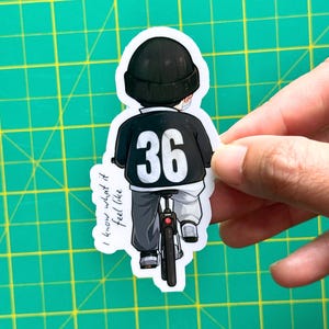 May include: A sticker featuring a cartoon illustration of a person riding a bicycle. The person is wearing a black beanie, a black jacket with the number 36, and gray pants. The text "I know what it feel like" is on the left side of the sticker.