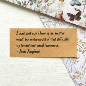May include: A brown card with a quote by Jeon Jungkook, a member of the South Korean boy band BTS. The quote reads: "I can't just say 'cheer up no matter what', but in the midst of that difficulty, try to find that small happiness."