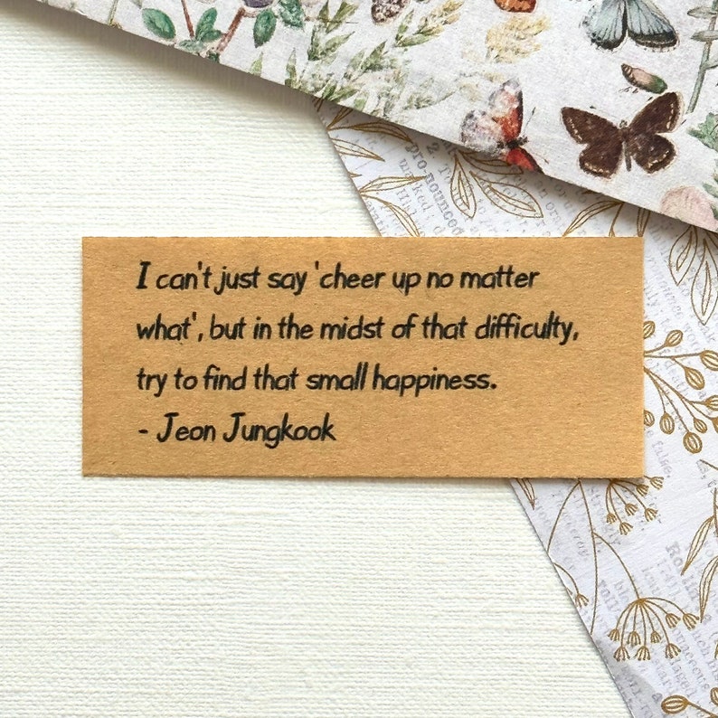 May include: A brown card with a quote by Jeon Jungkook, a member of the South Korean boy band BTS. The quote reads: "I can't just say 'cheer up no matter what', but in the midst of that difficulty, try to find that small happiness."