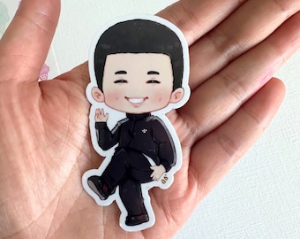 Pode incluir: Um adesivo de um personagem de desenho animado com cabelo preto, vestindo uma jaqueta preta com um logotipo branco, calças pretas e sapatos brancos. O personagem está sorrindo e acenando.
