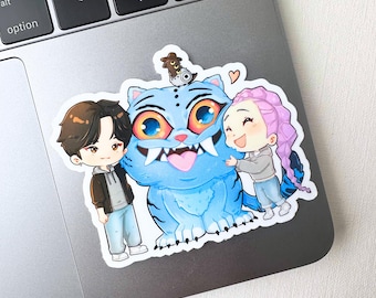 Könnte beinhalten: Ein Aufkleber mit einer blauen Cartoon-Katze mit großen Augen, scharfen Zähnen und einem kleinen Wesen auf dem Kopf. Zwei Cartoon-Figuren umarmen die Katze. Der Aufkleber befindet sich auf einem Laptop.