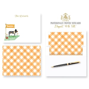 May include: A set of personalized notecards with a gingham pattern in orange and white. The notecards feature a black and white illustration of a dog with a Tennessee flag and a football. The text "The Warners" is printed in gold on the notecard. The text "Professionally Printed Notecards Elegant 110 lb. Felt" is printed in gold on the envelope.