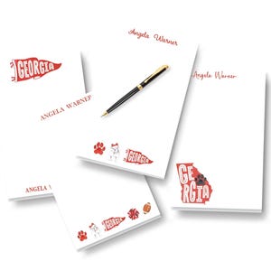 May include: A collection of white notepads with red accents. Each notepad features the name "Angela Warner" and various graphics, including a pennant with "Georgia" and a bulldog. A black and gold pen rests on one notepad.