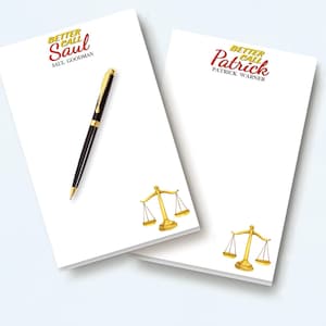 May include: Two white rectangular notepads featuring gold scales of justice at the bottom. One notepad displays the text Better Call Saul and Saul Goodman, while the other shows Better Call Patrick and Patrick Warner. A black and gold pen rests on top.