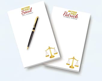 May include: Two white rectangular notepads featuring a gold and red graphic of a scale of justice. One notepad displays the text Better Call Saul Saul Goodman, the other Better Call Patrick Patrick Warner. A black and gold pen rests on the first pad.