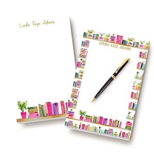 May include: Two white rectangular notepads featuring the name Linda Faye Adams in gold script. One notepad has a bottom border of watercolor books and plants, while the other has a full border of books and plants with a black and gold pen.