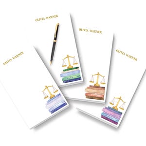 May include: Three white rectangular notepads with the name OLIVIA WARNER printed in gold at the top. Each notepad features a watercolor illustration of a gold scale of justice resting on a stack of books in varying colors. A black pen is included.