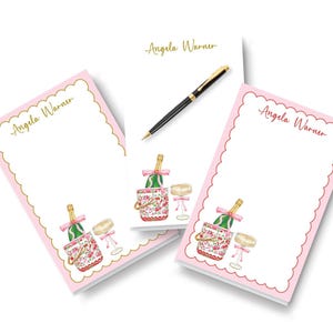 May include: Pink notepads with gold scalloped edges and the name "Angela Warner" in gold script. Each notepad features a watercolor illustration of a champagne bottle, ice bucket, and glass. A black pen rests on top.