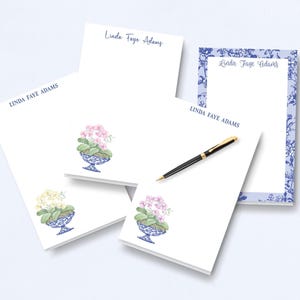 May include: White notepads with the name "Linda Faye Adams" in blue script. Some pads feature a watercolor illustration of a floral arrangement in a blue and white decorative bowl. A black pen with gold accents rests on one pad.