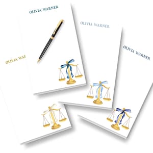 May include: Four white rectangular notepads featuring the printed name OLIVIA WARNER in various colors and a watercolor illustration of a gold scale of justice with a ribbon. A black and gold pen rests on the top notepad.