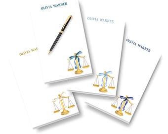May include: Four white rectangular notepads featuring the printed name OLIVIA WARNER at the top and a watercolor illustration of a gold scale of justice with a blue ribbon at the bottom. A black and gold ballpoint pen rests on one notepad.