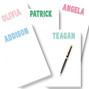 May include: Four white notepads with different names written in colorful outlined text. The names are Olivia, Addison, Patrick, and Angela. One notepad has a black pen on it and the name Teagan written in green outlined text.