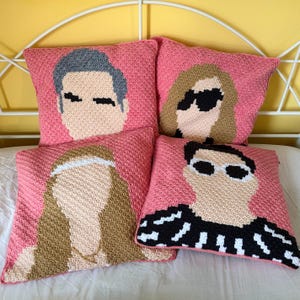 May include: Four pink crochet cushions featuring portraits of people. The portraits are created with various yarn colours, including black, white, tan, and grey. The cushions are square and appear to be handmade.