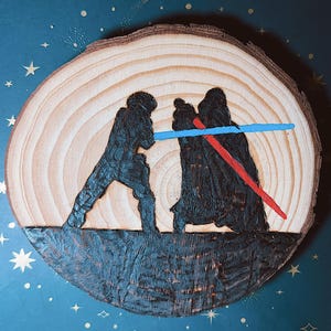 May include: A wood slice featuring a Star Wars scene with two figures engaged in a lightsaber duel. One figure wields a blue lightsaber, the other a red one. The artwork is burned into the wood, with a dark blue background.