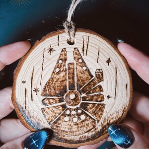 May include: A wooden, circular ornament featuring a pyrographed design of the Millennium Falcon starship. The ornament is made from a cross-section of a tree branch, with natural wood grain and bark edge. It has a twine loop for hanging.