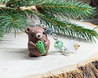 May include: A brown bear figurine holding a green fish, attached to a keychain with green and blue beads, and a white flower charm. The bear has black eyes and a tan snout. The keychain is displayed on a wooden surface with evergreen branches.