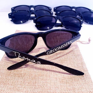 May include: Black sunglasses with the word "Groomsman" printed on one arm and "Thomas" printed on the other arm. The sunglasses are on a brown burlap bag.