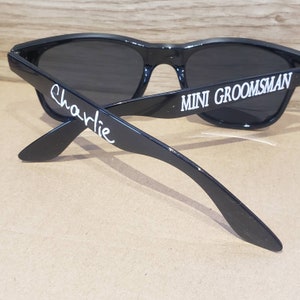 May include: Black sunglasses with the text "MINI GROOMSMAN" on one arm and "Charlie" on the other arm.