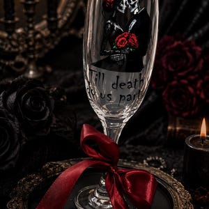 May include: A clear glass champagne flute featuring a graphic of a skeleton couple in formal attire holding red roses. The text "Till death do us part" is printed below the figures. A deep red satin ribbon is tied around the stem of the glass.