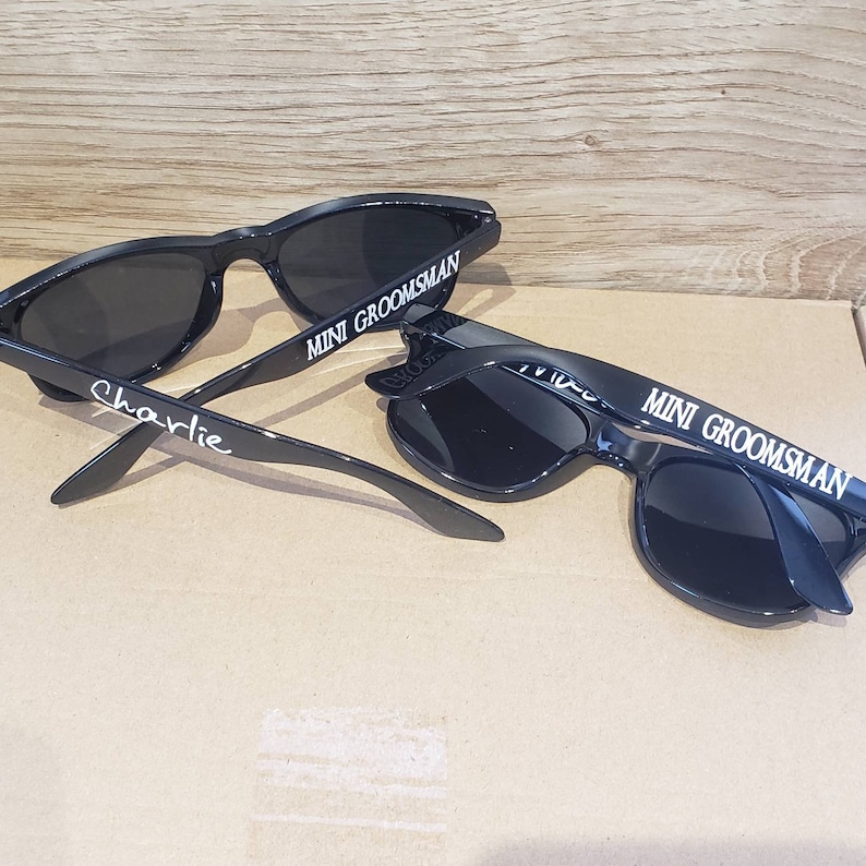May include: Two black sunglasses with the text "MINI GROOMSMAN" on the frames. One pair has the name "charlie" written on the arm.