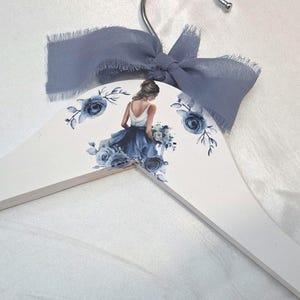 May include: White wooden clothes hanger decorated with a blue floral design and a watercolor image of a woman in a blue dress. A blue ribbon is tied at the top of the hanger.