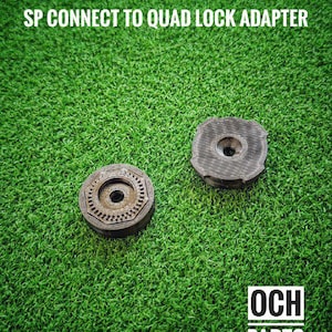Op de afbeelding: Twee ronde SP Connect to Quad Lock adapters met een centraal gat, op een groene ondergrond. De adapters zijn donker en getextureerd. De tekst "SP CONNECT TO QUAD LOCK ADAPTER" staat bovenaan.