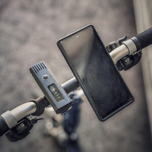 May include: A black smartphone is mounted on the handlebars of a black electric scooter. A black and gray device with a digital display is also mounted on the handlebars.