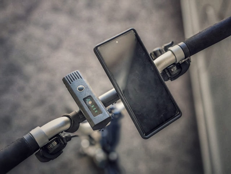 May include: A black smartphone is mounted on the handlebars of a black electric scooter. A black and gray device with a digital display is also mounted on the handlebars.