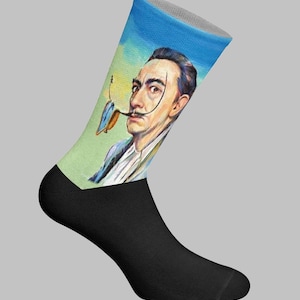 Peut inclure: Une paire de chaussettes noires et colorées représentant un portrait de Salvador Dalí avec sa moustache et sa pipe caractéristiques. Le haut de la chaussette affiche une peinture de Dalí sur un fond bleu et vert.