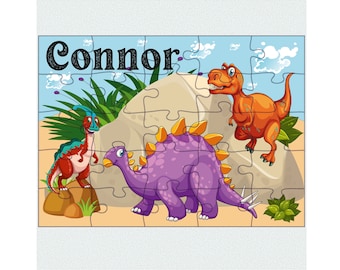 May include: A colorful jigsaw puzzle featuring three cartoon dinosaurs: a red and teal Parasaurolophus, a purple Stegosaurus, and an orange Tyrannosaurus Rex. The name "Connor" is printed in black at the top. The puzzle is set against a blue sky background.