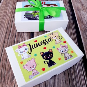 Puede incluir: Una caja de regalo blanca con una cinta verde y una ilustraci&oacute;n colorida de gatos y el nombre "Janessa" en la tapa. La caja viene con una caja m&aacute;s peque&ntilde;a separada con una ilustraci&oacute;n de cami&oacute;n.