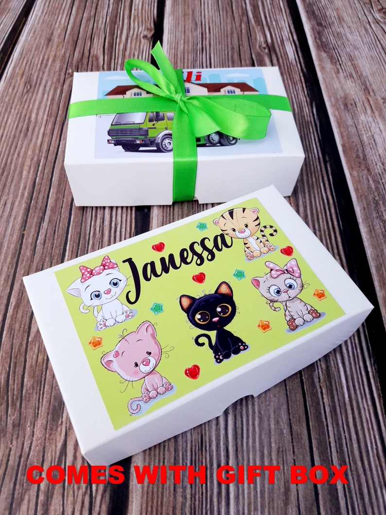 Puede incluir: Una caja de regalo blanca con una cinta verde y una ilustraci&oacute;n colorida de gatos y el nombre "Janessa" en la tapa. La caja viene con una caja m&aacute;s peque&ntilde;a separada con una ilustraci&oacute;n de cami&oacute;n.